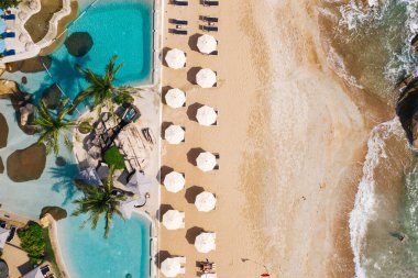 Açık havuzu ve güneş pansiyonları olan ilk hat plaj otelinin havadan görünüşü. Yaz tatillerinin kuş bakışı manzarası, turkuaz deniz suyu ve temiz kum kıyısındaki şemsiyeler.