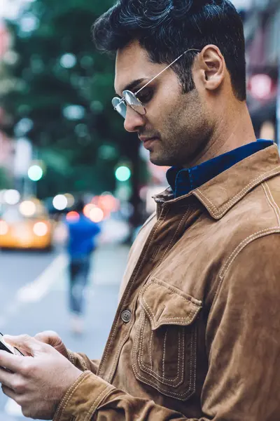 Sakin, yakışıklı, ceketli ve gözlüklü yetişkin bir adamın gündüz şeridinin yanında dururken akıllı telefon kullanmasının yan görüntüsü.