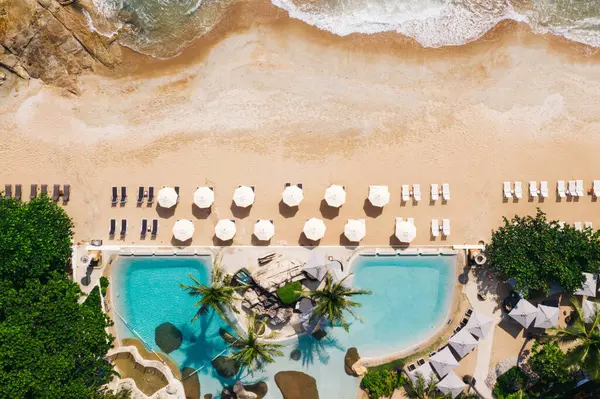 Açık havuzu ve güneş pansiyonları olan ilk hat plaj otelinin havadan görünüşü. Yaz tatillerinin kuş bakışı manzarası, turkuaz deniz suyu ve temiz kum kıyısındaki şemsiyeler.