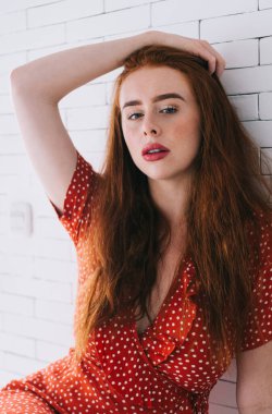 Kırmızı benekli elbiseli, kırmızı saçlı, dalgın bir kadın el üstünde oturuyor ve kameraya bakarken beyaz tuğla duvarın yanında dinleniyor.