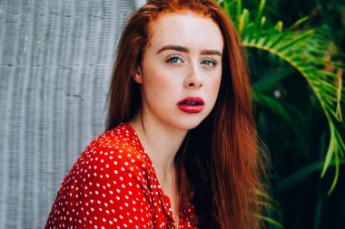 Kırmızı benekli elbiseli, düşünceli ve odaklanmış kızıl saçlı kadın bitkilerin yanındaki hasır sandalyede oturuyor ve kameraya bakıyor.