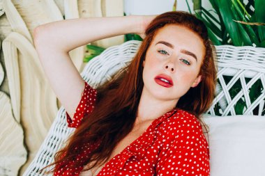 Kırmızı noktalı elbiseli şehvetli kızıl saçlı kadın hasır sandalyeye uzanmış yeşil tropikal bitkilerin arka planındaki kameraya bakıyor.