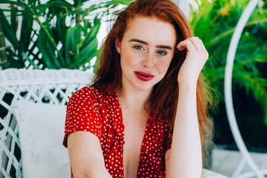 Kırmızı benekli kırmızı saçlı kadın beyaz koltukta oturuyor ve saçlarına dokunurken gülümsüyor ve kameraya bakıyor.