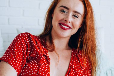 Puantiyeli akıllı telefonlu mutlu genç bayan beyaz duvara bakarken bacak bacak üstüne atmış bir şekilde yatağa uzanıyor.