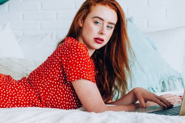 Laptoplu, benekli elbisesi beyaz yatakta yatıp kameraya bakan kızıl saçlı genç bir kadın.