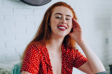 Güzel, kızıl saçlı, akıllı telefonlu bir kadın kafasına dokunuyor ve yatağında kameraya bakarken bacak bacak üstüne atıyor.