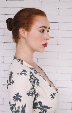 Beyaz tuğla duvarın önünde duran, güzel elbiseli, kızıl saçlı, çekici bir kadının yan görüntüsü.