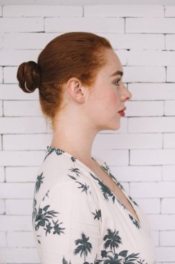 Beyaz tuğla duvarın önünde duran, güzel elbiseli, düşünceli, genç, düşünceli, kızıl bir kadının yan görünüşü.