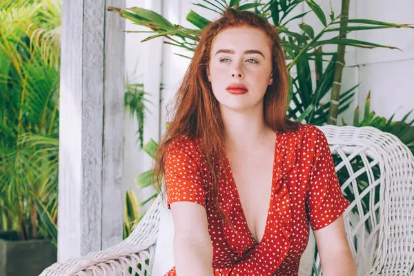 Güzel, düşünceli, kızıl saçlı, sıradan kırmızı noktalı elbiseli, kırmızı rujlu, evde cep telefonuyla dinlenen ve gözlerini çeviren bir kadın.