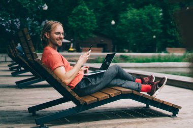 Modern dizüstü bilgisayarlı, gülümseyen ve akıllı telefonlu mutlu adamın yan görüntüsü parktaki gölün kenarında uzanırken.