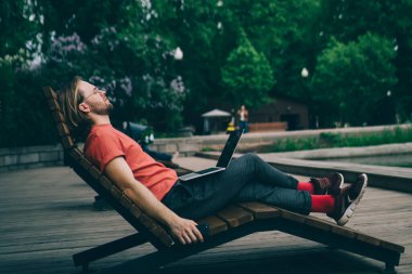 Modern dizüstü bilgisayarlı bir adamın, parktaki göl kıyısındaki iskelede serbest iş yaparken dinlenmesi.