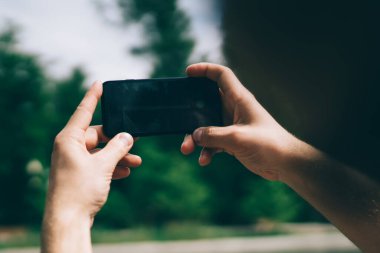 Tanımlanamayan adam, güneşli yaz gününde yeşil parkta dinlenirken bulanık göletin fotoğrafını çekmek için modern akıllı telefonu kullanıyor.