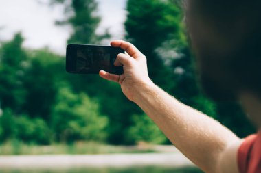 Tanımlanamayan bir adam, yazın parkta gölün bulanık arka planında dururken modern akıllı telefon kullanarak selfie çekiyor.