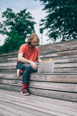 Sakallı modern hippi gözlüklü kırmızı çoraplı, uzun saçlı, kırmızı tişörtlü, dizüstü bilgisayarda gezinen ahşap bankta oturuyor.