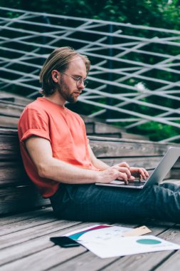 Gündüz vakti elinde kağıtlar ve akıllı telefonla tahta zeminde otururken dizüstü bilgisayarda çalışan genç adam.