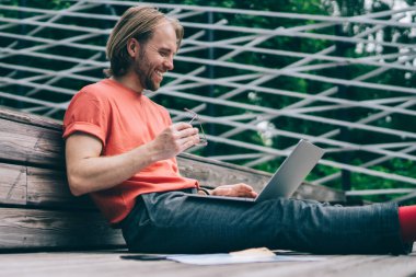 Parktaki ahşap platformda oturan dizüstü bilgisayarla gülen neşeli, genç bir hippi.