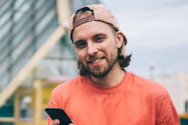 Günlük kıyafetleri ve şapkası olan neşeli erkek akıllı telefonu sosyal ağda gezinmek için kullanıyor. Şehir sokaklarında kameralara bakıyor.