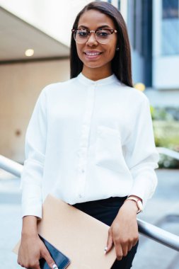 Başarılı koyu tenli ofis çalışanı yarım uzunluk portre klasör ile smartphone aygıt finans şirketi building yakınındaki kentsel ortamda ayakta elinde tutan şık beyaz gömlek giymiş