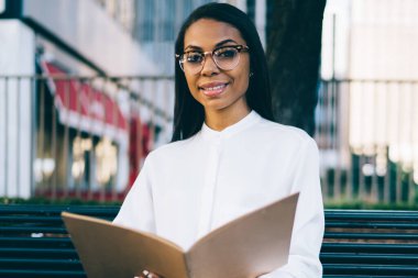 Portre pozitif Afrikalı-Amerikalı kadın çalışanın açık havada kentsel bankta belirleyen ve finansal belgeler klasöründe kontrol otururken kameraya gülümseyen şık beyaz gömlek giymiş