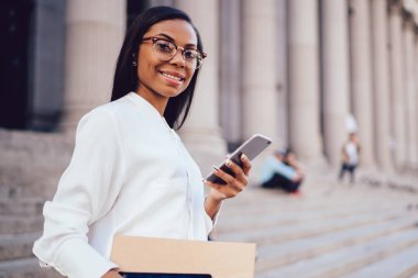 Başarılı büyüleyici işkadını klasör ile dokunmatik yüzey elinde tutan ve smartphone ayakta şehir merkezinde kentsel ortamda profil güncelleştirme sırasında kameraya gülümseyen koyu tenli portresi