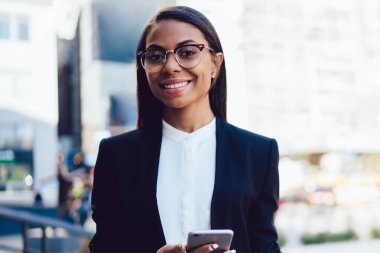 Formal giyim klasör ve smartphone aygıt kentsel ortamda şehir merkezinde duran elinde tutan yarım uzunluk portre avukat Fakültesi neşeli Afrikalı-Amerikalı öğrencinin giymiş