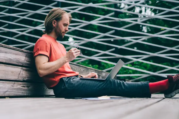 Düşük açılı, rahat, gözlüklü, uzaktan kumandayla çalışan, parktaki ahşap platformda oturan genç bir adam.