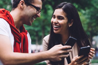 Gözlüklü beyaz gömlekli erkek arkadaş ve modaya uygun giyinmiş kız arkadaş hem akıllı telefon kullanıyor hem de arka planda bulanık bir şekilde gülüyor.