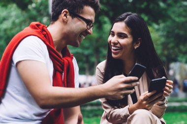 Gözlüklü beyaz gömlekli erkek arkadaş ve modaya uygun giyinmiş kız arkadaş hem akıllı telefon kullanıyor hem de arka planda bulanık bir şekilde gülüyor.