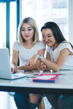 Sıradan giyinmiş, gözlüklü, neşeli, etnik tebessümlü bir kadın, kafedeki sarışınla gevşeyen dizüstü bilgisayarın ekranını gösteren bir video izliyor.