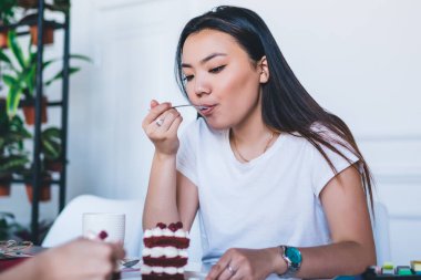 Beyaz tişörtlü çeşitli genç kadınlar kahve fincanları içip arka planda saksılarda bitkiler olan modern beyaz kafeteryadaki kekleri tadıyorlar.