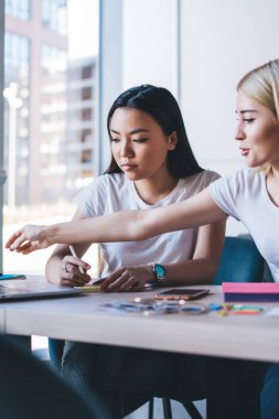 Beyaz tişörtlü çok ırklı kadınlar laptopla çalışıyor ve modern beyaz ofisteki insanlarla masaya oturarak notlar alıyorlar.