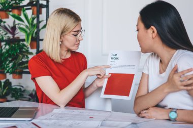 Yoğunlaşmış gözlüklü genç bayan modern çalışma alanında gündelik giysiler içinde etnik kadınla ortak girişim yaratan belgeye işaret ediyor.