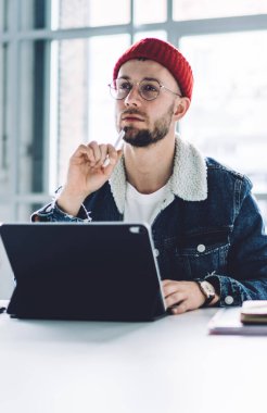 Genç, düşünceli, sakallı, gözlüklü, sıcak elbiseli, parlak odalı, pencereli, dizüstü bilgisayarlı biri.