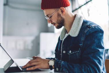 Tarz sahibi, sakallı, gözlüklü ve şapka takan genç bir erkek öğrencinin beyaz odadaki tablete mesaj yazışının yan görüntüsü bulanık arka planda.