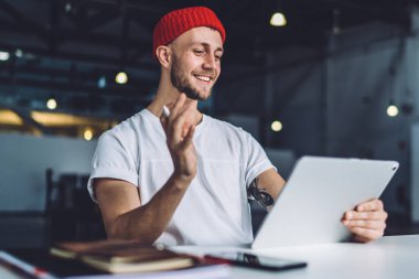 Kırmızı bereli neşeli ve hevesli adam modern geniş ofisteki masada Stylus 'la çalışırken tablet üzerinde video görüşmesi yapıyor.