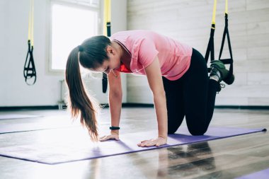 Pembe tişört ve siyah tayt giyen genç ve çekici bayan sporcunun yan görüntüsü stüdyodaki havacılık dersi sırasında mavi spor paspasın üzerinde çalışıyor.