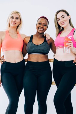 Aktif giyimli bir grup iyimser ve enerjik spor kadın beyaz spor salonunda kameraya sarılıp bakarken