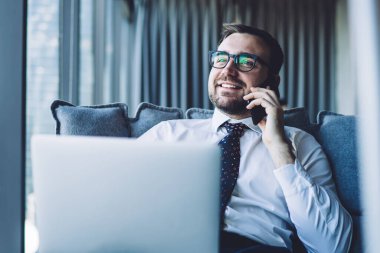 Optik gözlüklü mutlu işadamı bilgisayardaki online iş sırasında şirket bilgilerini tartışmak için akıllı telefon konuşması yapıyor, cep telefonu cihazıyla konuşan neşeli CEO.