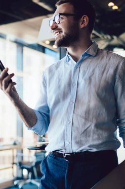 Cep telefonu cihazını tutan iyi görünümlü bir ofis çalışanı çalışanlarla cep telefonu iletişimi sırasında başka tarafa bakıyor ve gülümsüyor, resmi giyinmiş mutlu bir işadamı akıllı telefon teknolojisinde 4G internet kullanıyor.
