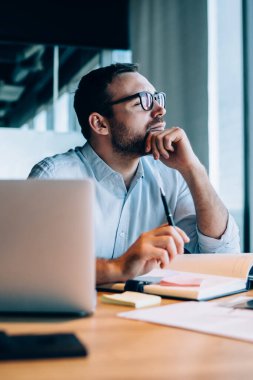 Gözlüklü entelektüel sakallı bir adam mavi gömlekli pencereden dışarı bakıyor. Not defterine not alıyor. Masada dizüstü bilgisayarla oturuyor.