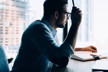 Genç, gözlüklü, sakallı, pencereye bakan, dizüstü bilgisayar ve defterle çalışırken kalemle elini kaldıran adamın yan görüntüsü.