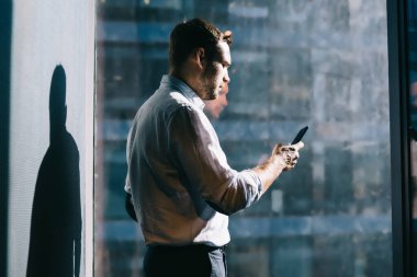 Resmi tişörtlü, ciddi bir erkek. Akıllı telefonuyla mesajlaşıyor. Ofis odasının camına yansıyan büyük pencerenin yanında duruyor.