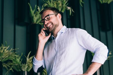 Modern ofisteki saksı bitkileriyle dekore edilmiş duvarın yanında dururken akıllı telefondan gülümseyen ve konuşan mutlu erkek yöneticinin alçak açısı.