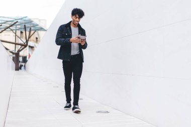 Modern stil güler yüzlü, neşeli, esmer bir adam. Gündüz vakti sokak tüneli boyunca yürürken tablet üzerinde geziniyor.