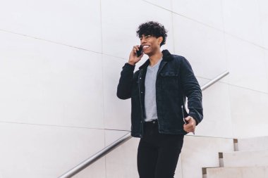 Modern stil, mutlu, gülümseyen, Afro-Amerikalı bir adam. Günlük kıyafetleriyle telefonda konuşurken gün ışığında şehir merdivenlerinde yürürken.