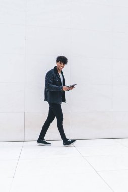 Modern stil, güler yüzlü, kendinden emin, Afro-Amerikan bir adamın gün ışığında şehir caddesinde yürürken tablet üzerinde gezinmesinin yan görüntüsü.