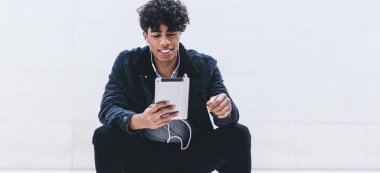 Afro-Amerikalı, kıvırcık saçlı, gülümseyen ve beyaz merdivenlerde oturan, tablette mesajlaşan ve kulaklıkla beyaz duvarda müzik dinleyen genç bir erkek.