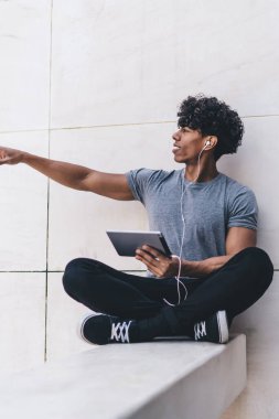 Neşeli, mutlu, Afro-Amerikalı, sıradan kıyafetler giymiş, tablet ve kulaklıklarla bacak bacak bacak üstüne atmış, açık havada kollarını gösteren yakışıklı bir erkek.