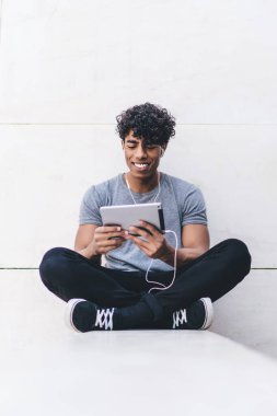 Gülümseyen, yakışıklı, sıradan giyinmiş, bacak bacak bacak üstüne atmış, dışarıda kulaklıklarla video izleyen siyah bir erkek.