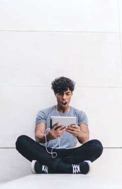 Şok olmuş genç, sıradan kıyafetlerle yakışıklı bir erkek bacak bacak bacak üstüne atıyor ve dışarıda kulaklıklarla video seyrediyor.